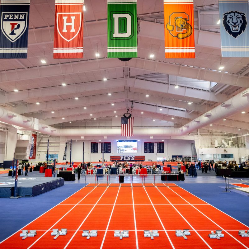 Ivy league school banners hang above an orange track with spectators enjoying the Ott Center indoor track facility ribbon cutting ceremony.